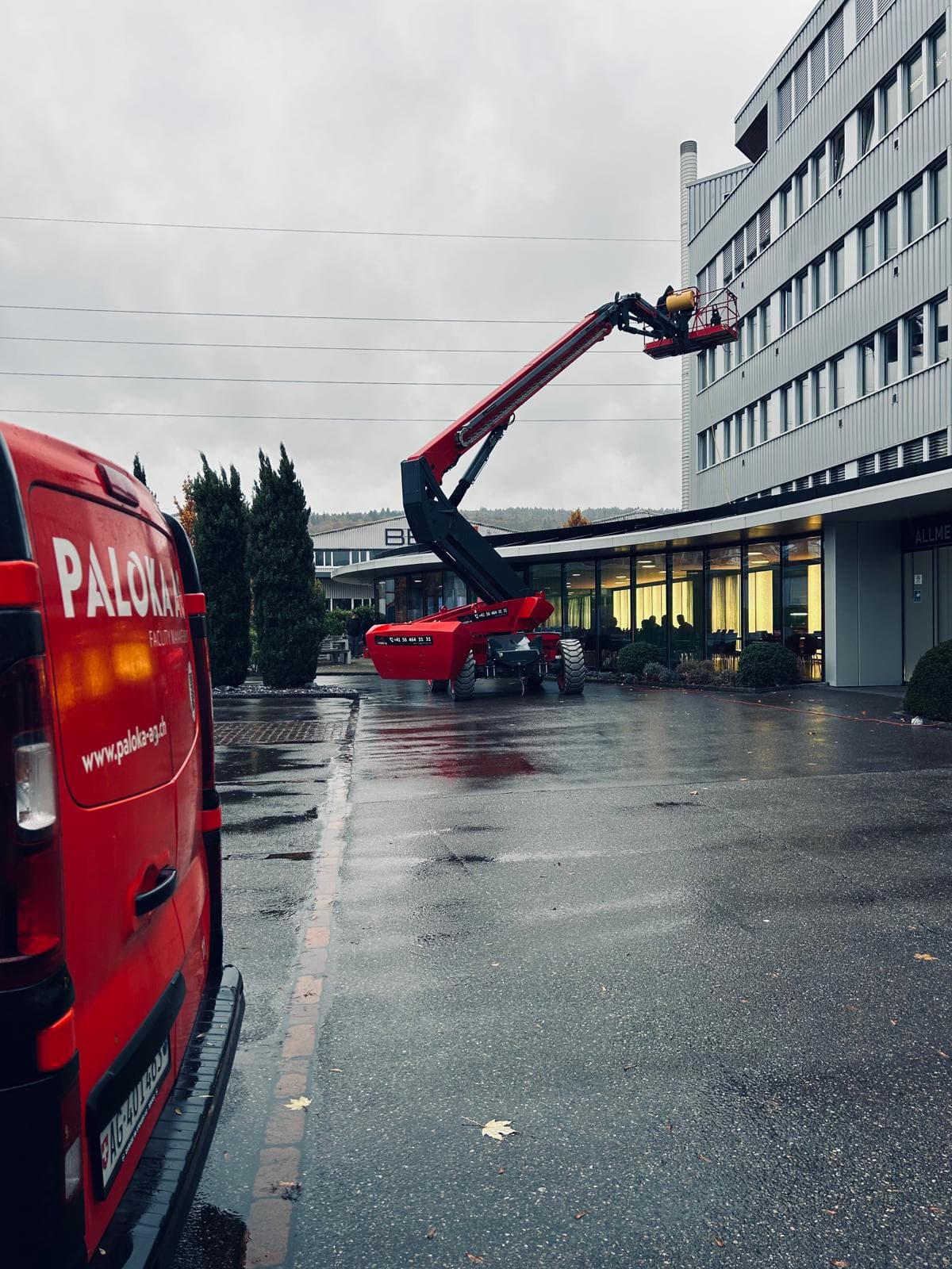 Eine rote Hebebühne an der Fassade eines mehrgeschossigen Gebäudes, mit einem Paloka-Fahrzeug im Vordergrund.