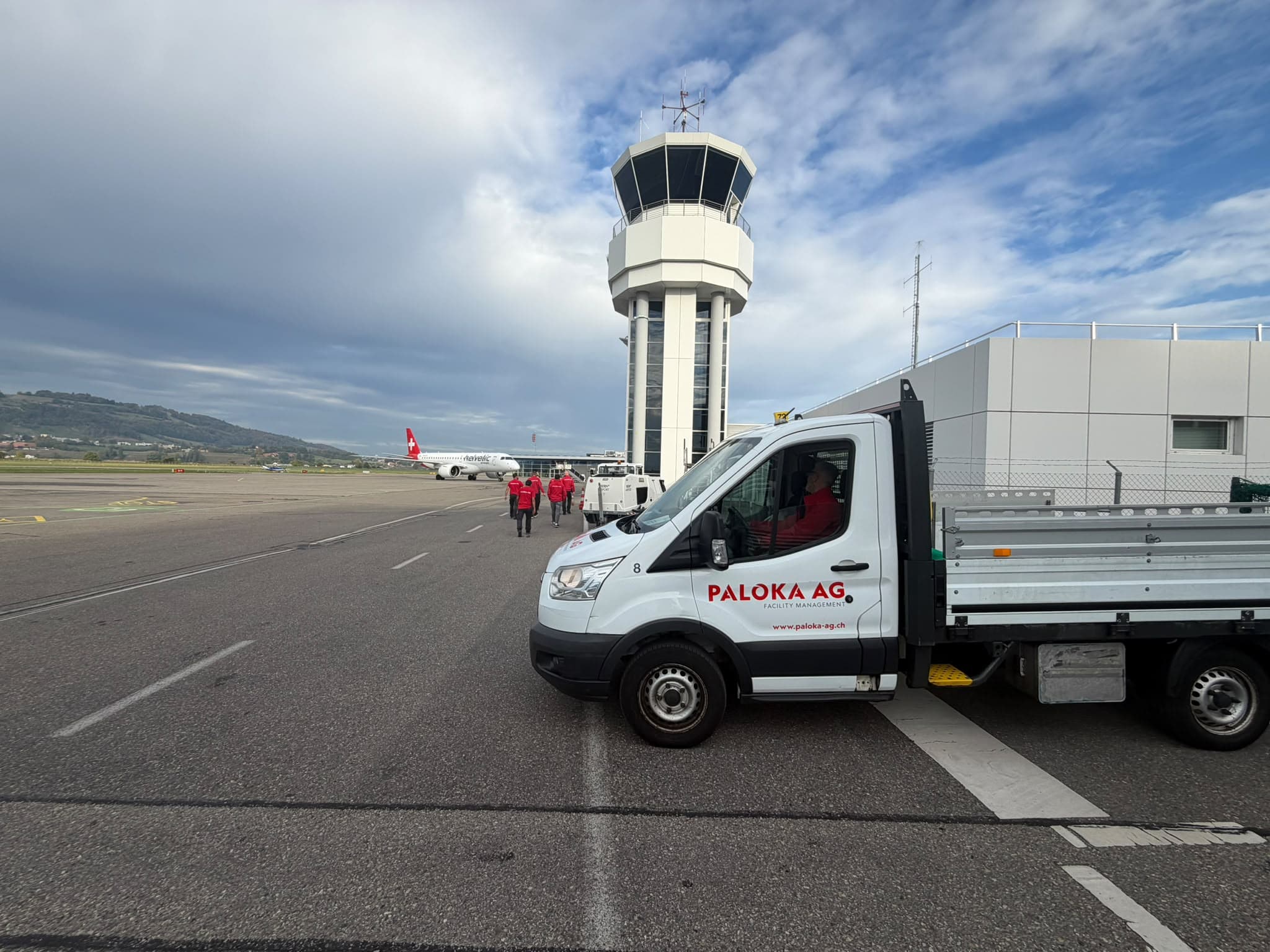Paloka-Fahrzeug und Team auf einem Flughafenvorfeld, mit Kontrollturm und Flugzeug im Hintergrund.