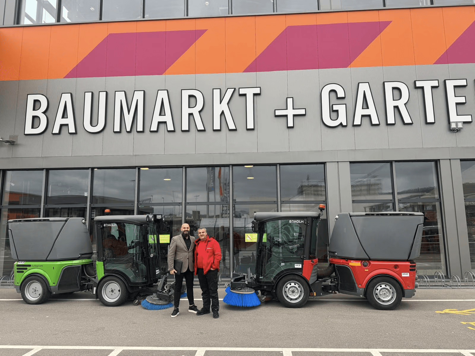 Zwei Personen zwischen einer grünen und einer roten Kehrmaschine vor einem Baumarkt.