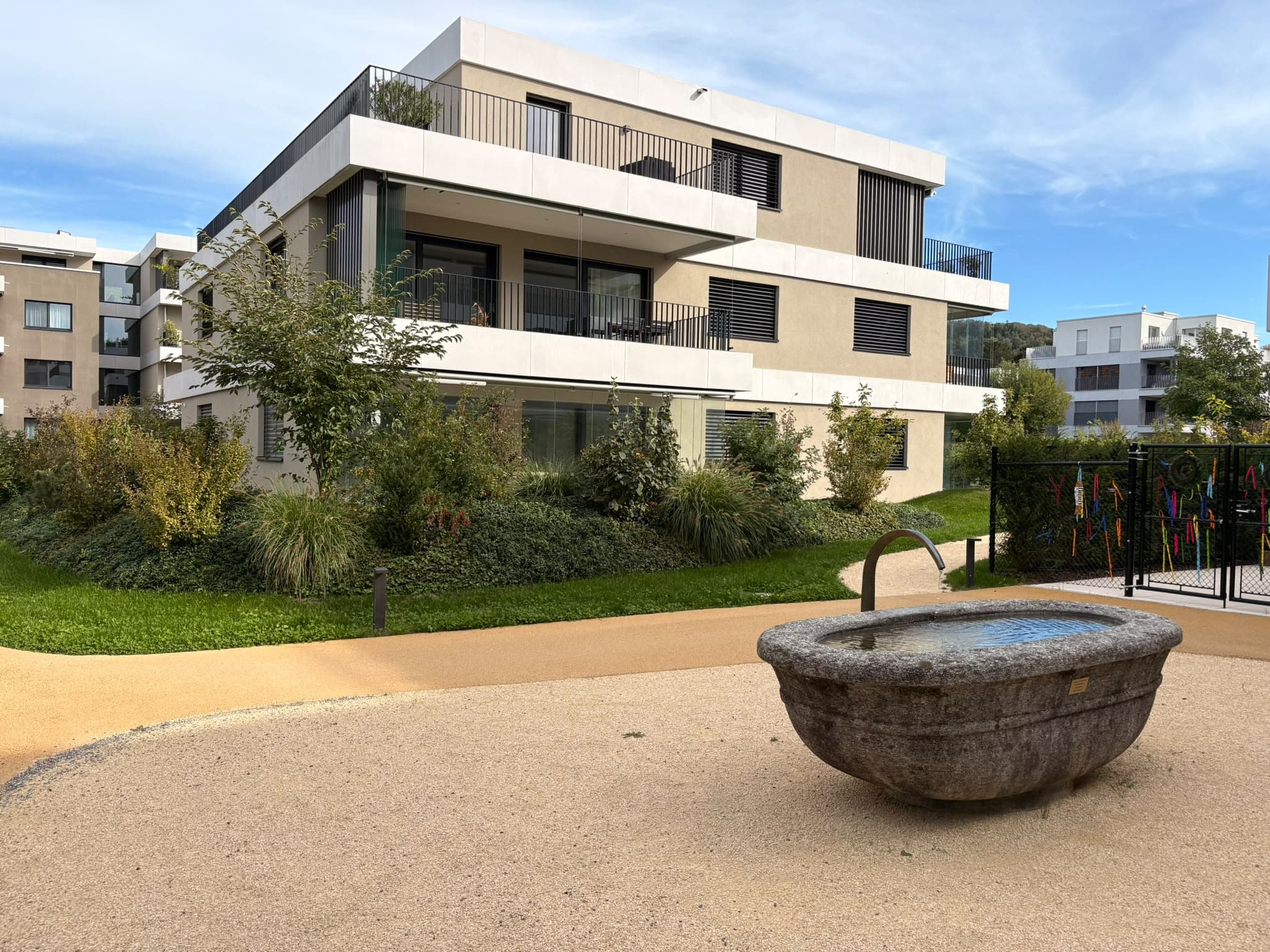 Wohnhaus in Eglisau mit breiten Balkonen, begrünten Aussenflächen und einem steinernen Brunnen im Vordergrund.