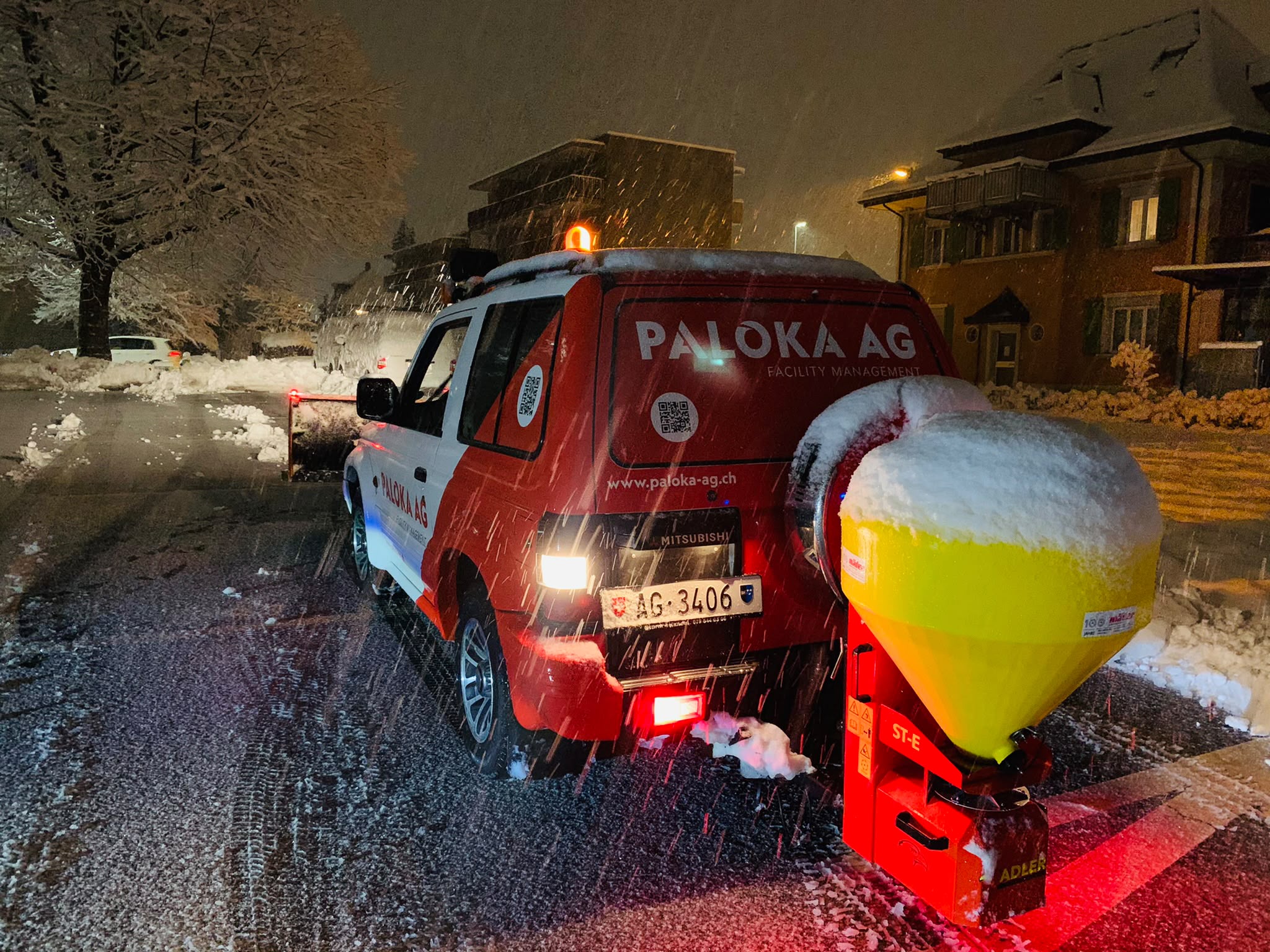 Paloka-Winterdienstfahrzeug mit Schneepflug, gelbem Streugerät und eingeschaltetem Warnlicht bei nächtlichem Schneefall.